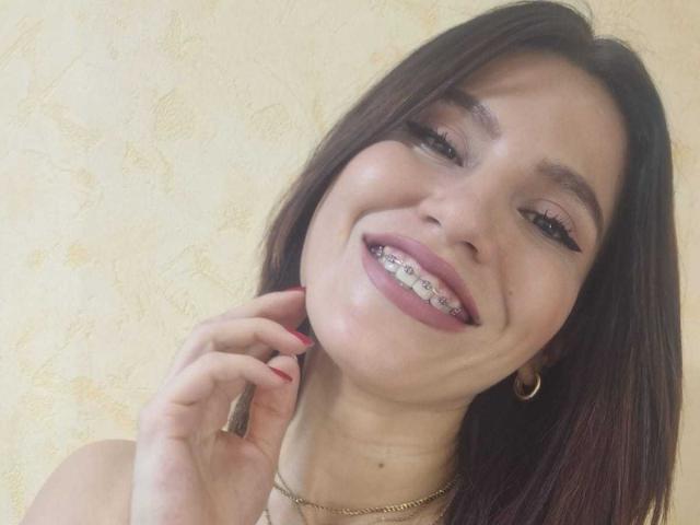 AmallaVega has a smile with her teeth showing, brown hair, red nails, gold hoop earrings, and a gold necklace. She's touching her neck with her right hand.