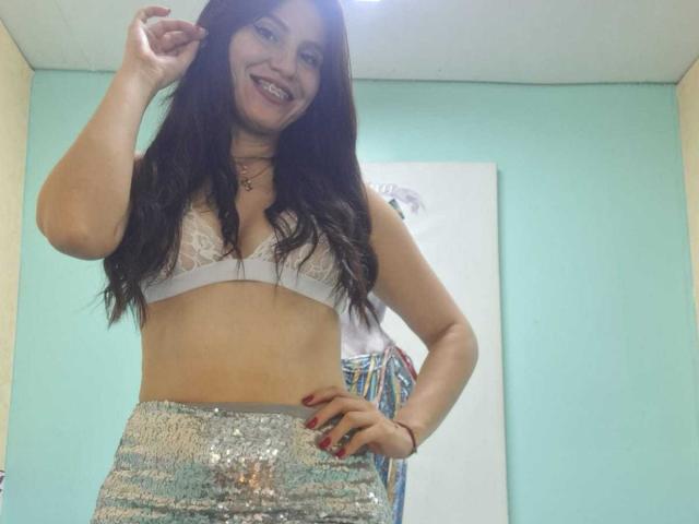 AmallaVega with long dark hair, smiling, wearing a white lace bra and silver sequined skirt, hand in hair, red nails, light blue wall, and a small painting in background.