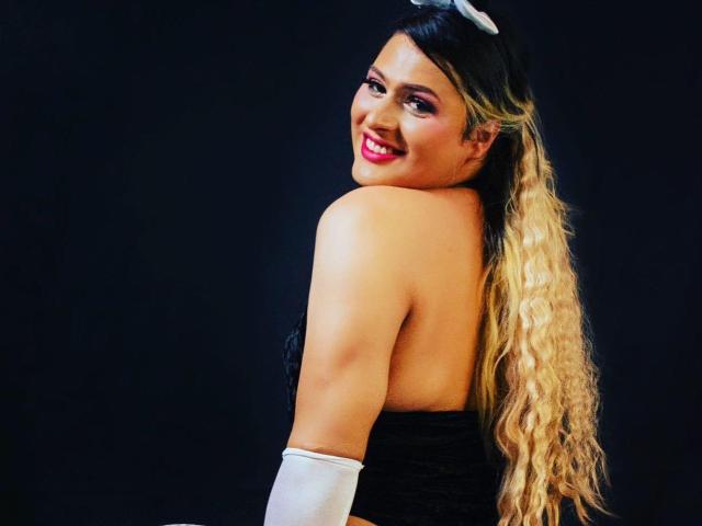 DreamsBlondeBebe with long, wavy blonde hair, smiling at the camera, showing her bare back and white arm glove, wearing a black strapless dress.