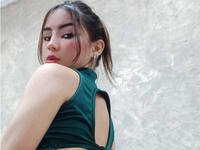 AshleyMiler with pouty red lips, green velvet top, back cutout, brown hair in ponytail, looking over shoulder, fair skin, earrings, slight blush on cheeks.