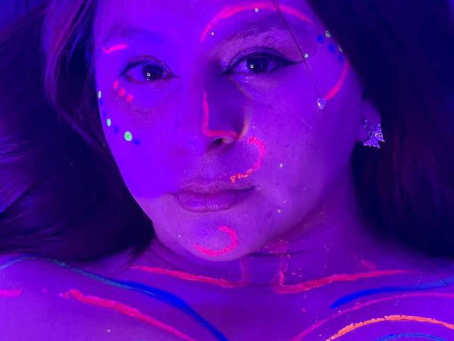 EmaJames's face and upper chest are covered in glowing neon pink and blue patterns, with purple lighting, her eyes looking forward, wearing a butterfly earring, and lips slightly parted.