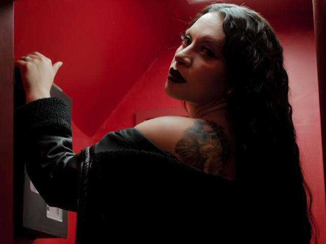 KatalinaJohnson with dark lipstick, black off-shoulder sweater, long wavy hair, tattoo on right shoulder, looking back, hand on red wall, red lighting, slightly turned head.