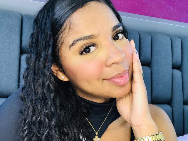 LisaMendez with curly black hair, brown eyes, and a cute smile, touching her cheek, wearing a black top and gold necklace, showing her gold watch.