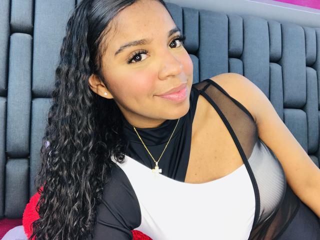 LisaMendez with curly black hair, brown eyes, and a slight smile, wearing a black and white top, showing her tits, sitting on a gray cushioned surface.