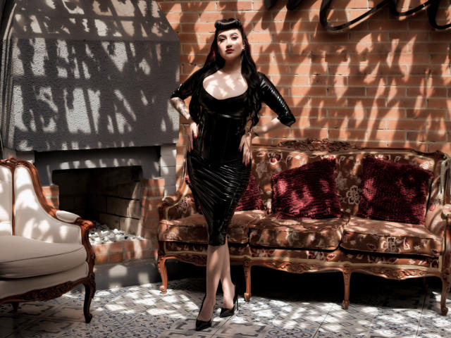 EmmaRosseti stands in a black latex dress with cleavage, black heels, and a hat, hands on hips, in a sun-dappled room with a vintage couch and red pillows.