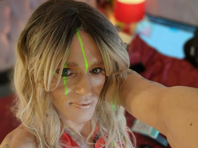 Eine Frau mit blonden Haaren und grünen Make-up-Linien im Gesicht, die in die Kamera lächelt und ein Selfie macht, trägt ein rotes Hemd, hat welliges Haar und leichte Sommersprossen. Im unscharfen Hintergrund ist eine rote Lampe zu sehen, und es handelt sich um ein Schlafzimmer..