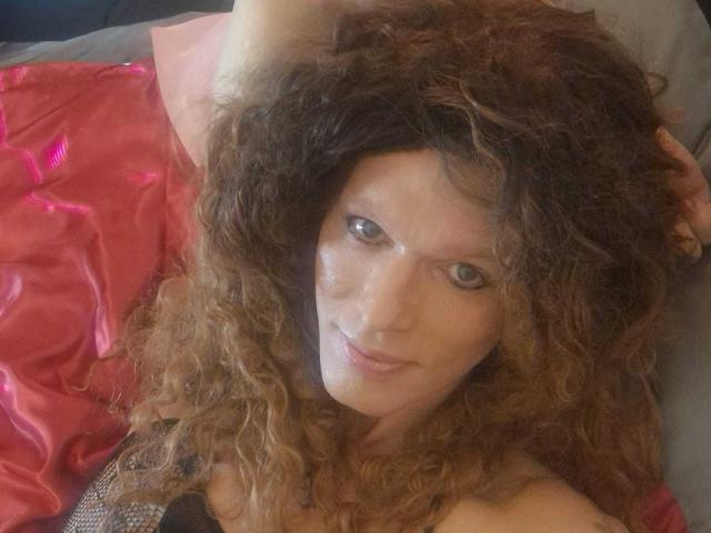 Cherris has curly brown hair, a shiny face, and a slight smile, lying on a red satin sheet, looking up with blue eyes. Her hand rests on her head.