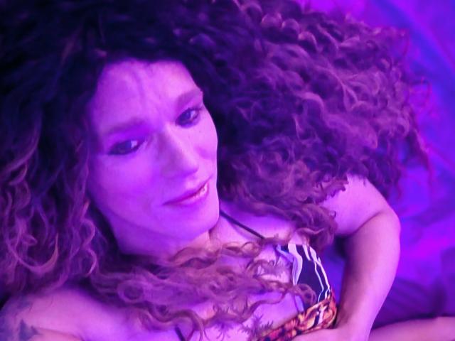 Eine Frau namens Cherris mit lockigem, braunem Haar, einem violett beleuchteten Gesicht und einem farbenfrohen Oberteil, die mit einem leichten Lächeln nach oben schaut, die Arme verschränkt. Ihre Brüste sind teilweise sichtbar..