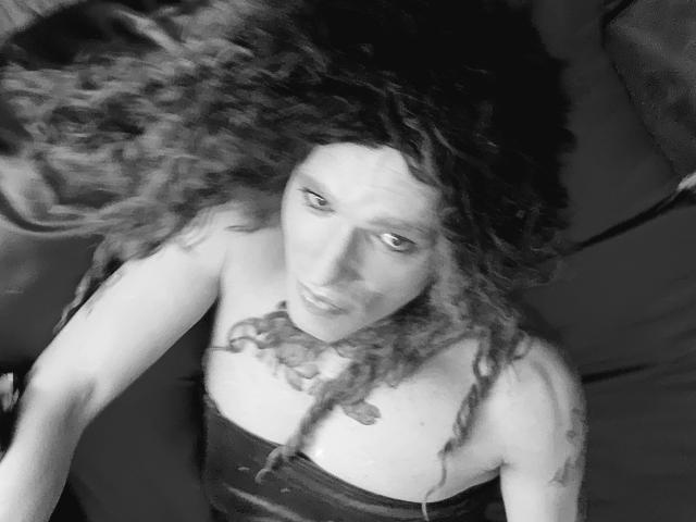 Photo en noir et blanc de Cherris, avec des cheveux bouclés, regardant vers le haut, portant un haut sans bretelles, montrant de petites seins et un tatouage sur la poitrine. Cherris a une expression sérieuse..