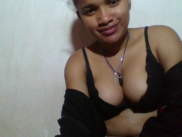 HoneyLie with dark skin and curly hair, wearing black lace bra, showing her big tits, black off-shoulder top, and necklace, smiling at the camera.