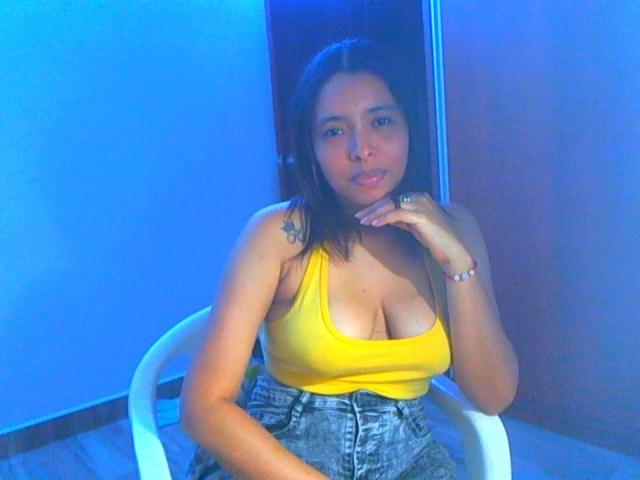 AnnaBellaStar69 with medium tits in a yellow tank top, sitting on a white chair, wearing high-waisted jeans, blue lighting, looking at camera, right hand near chin.