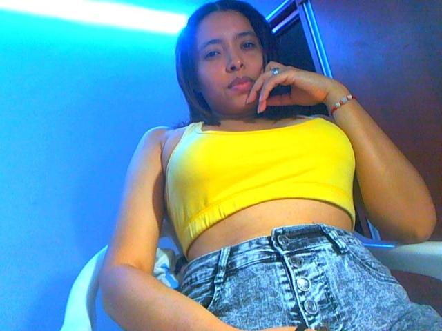 AnnaBellaStar69 with pouty lips, wearing a yellow crop top and high-waisted, button-down denim shorts, has her finger near her mouth, under blue neon light.