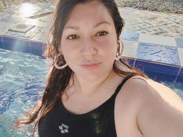 Sakoto Dika, con i capelli castani bagnati, indossa un costume intero nero e mostra il seno mentre è in piscina, con orecchini a cerchio rosa, ha una carnagione chiara e un leggero sorriso..