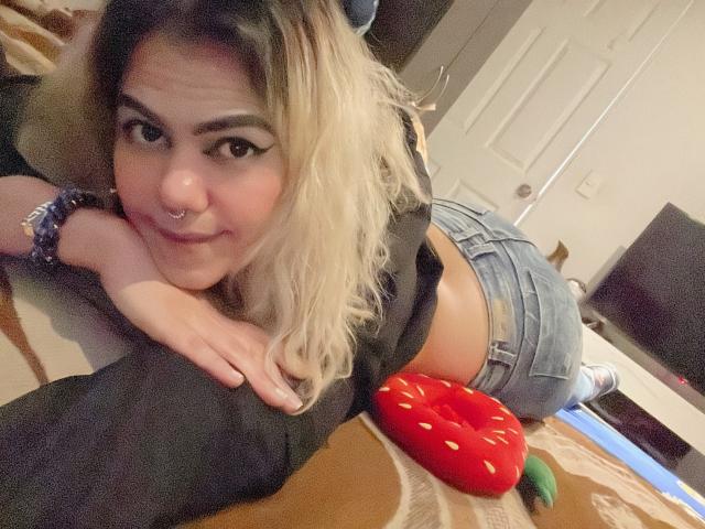 TarahDustin, with blonde hair and nose ring, lying on her stomach, showing her ass in denim shorts, smiling at the camera, wearing a black top, and a red donut toy.