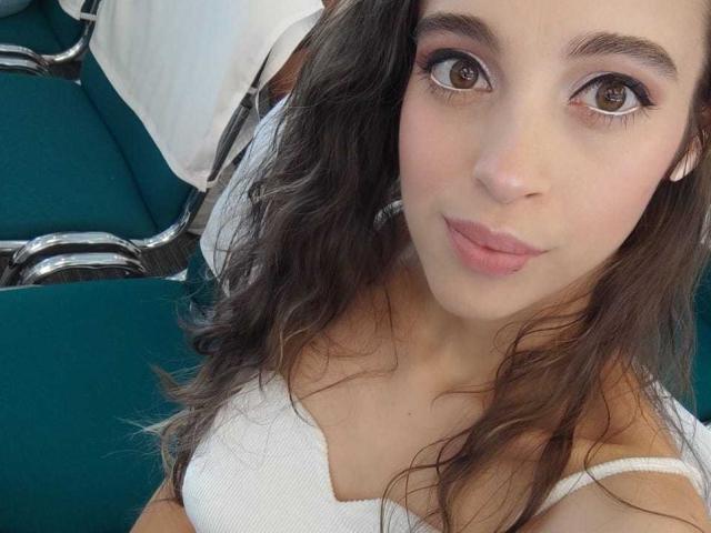 VioletaHade has brown eyes, wavy dark brown hair, and pink lips, wearing a white bra, sitting in a green chair with metal armrests.