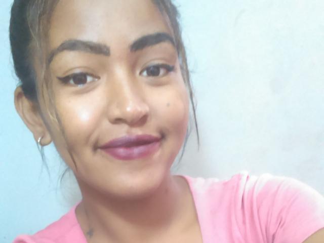 Syane's gezicht met donkere wenkbrauwen, een kleine neus en roze lippenstift. Ze glimlacht met een lichte grijns, draagt een roze shirt en heeft een kleine tatoeage op haar rechterhals..