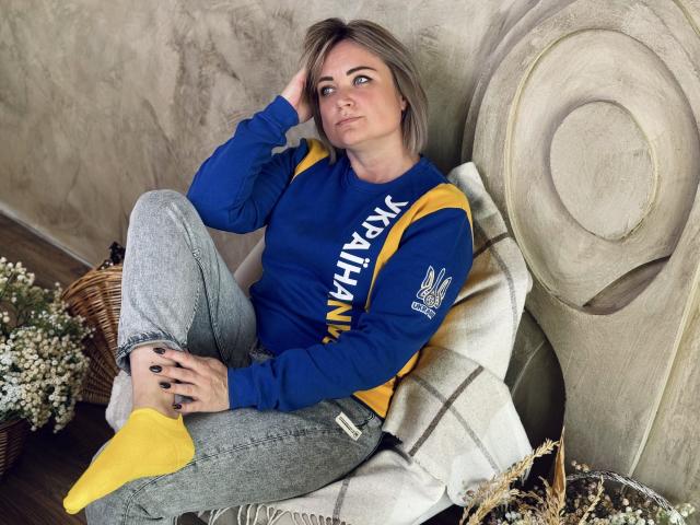 SnezhanaRells with short blonde hair, blue and yellow NY Yankees sweatshirt, grey jeans, yellow socks, black nails, and hand in hair, sitting on a white cushioned chair.