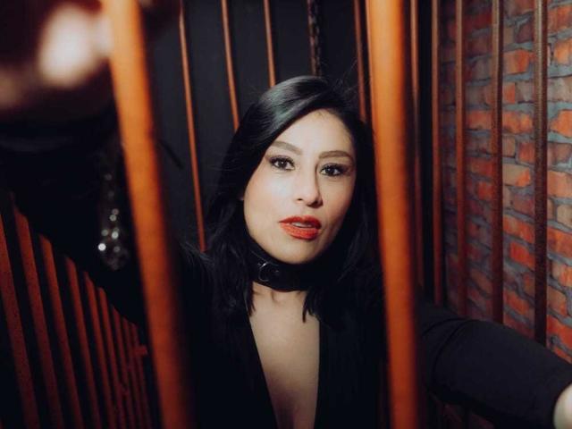 VioletaWhitee with black hair, red lipstick, and a black collar, looking through orange bars, has a sexy, confident expression, and is wearing a low-cut black top.