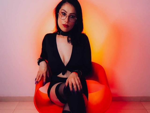 VioletaWhitee with glasses, black choker, deep cleavage, black top, black thigh-highs, sitting on orange chair, red-orange light, looking at camera, purple nails, white tile floor.
