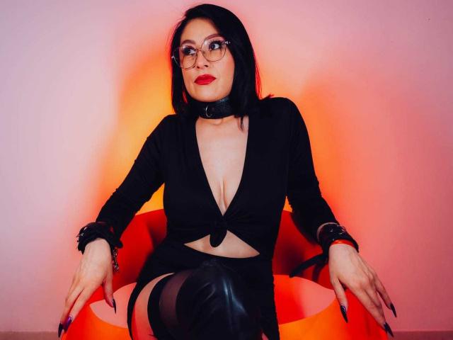VioletaWhitee with black bob, glasses, red lipstick, black tied top, cleavage, black cuffs, leather pants, sitting on orange chair, hands on thighs, red-orange light.