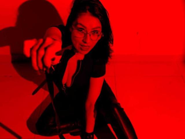 VioletaWhitee with red light, pointing forward, black dress, glasses, and a zipper, crouching, shadow on floor, intense gaze, black hair, wristband, and a leash in her hand.