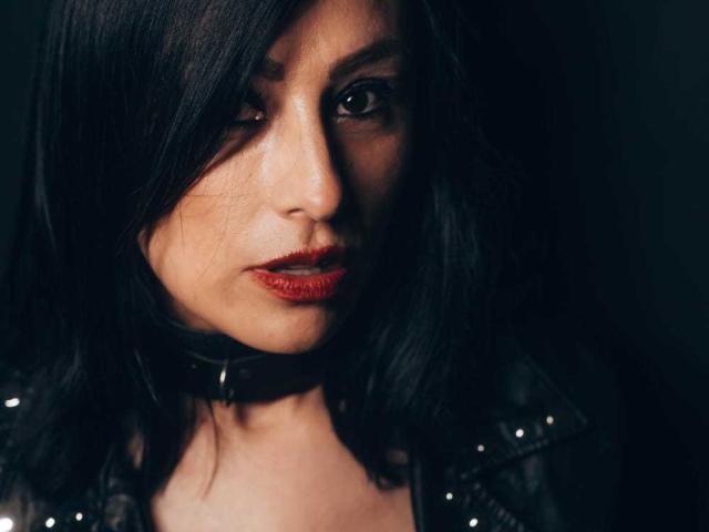 VioletaWhitee with dark, shoulder-length hair, red lipstick, and a black leather choker, looking intensely at the camera, wearing a studded leather jacket.
