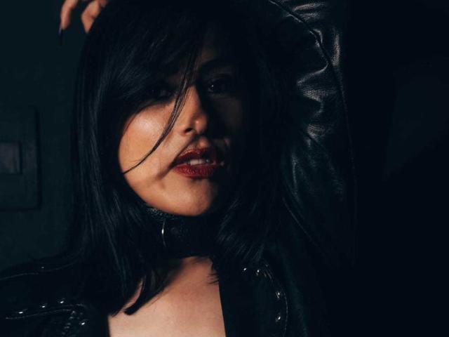 VioletaWhitee with black hair, dark leather jacket, and red lipstick, her hand in her hair, showing off her glossy red lips and slight smirk. Her tits are partially visible.
