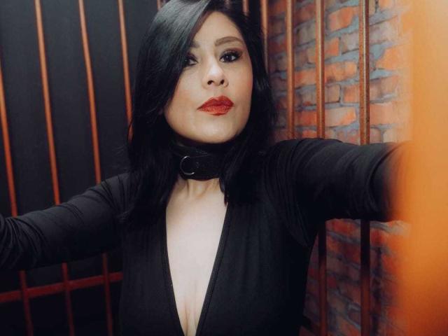 VioletaWhitee with black hair, red lipstick, and a deep V-neck black top, wearing a black choker with a ring, arms out, looking at the camera. Boobs visible.