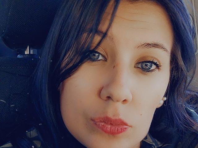HotBebedux has blue eyes, dark straight hair, light skin, small nose, and pouty red lips. She's making a puckered face in a car with a black headrest.