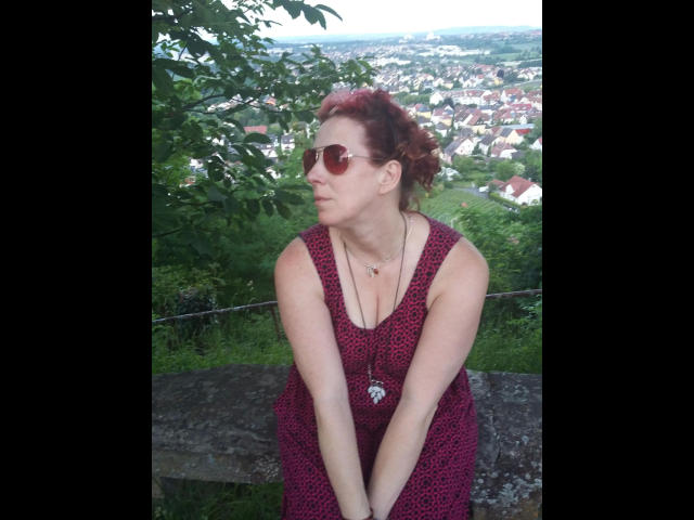 MissImmergeil76-hot with red hair, sunglasses, and a red leopard print dress, sitting on a rock, looking to the side, arms resting on knees, small tits visible, necklace.