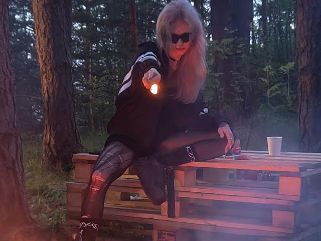 ExcitedBlizzard with blonde hair, sunglasses, black hoodie, ripped black leggings, and boots, sitting on wooden table, holding flashlight, with white cup and cigarette on table.