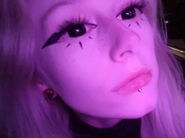 ExcitedBlizzard's face, purple-lit, with long eyelashes, black winged eyeliner, freckles, and a lip piercing. She's got pinkish skin and red ear studs.