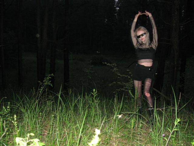 ExcitedBlizzard stands in a dark forest, arms raised, wearing black lingerie, fishnet stockings, and sunglasses, with grass in the foreground and tall trees in the background.