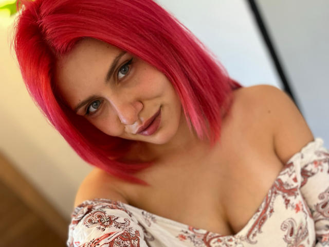 SophieReyX with pink hair, blue eyes, and a cute smile, wearing an off-shoulder patterned top, showing her tits. She has a slight nose and a sexy look.