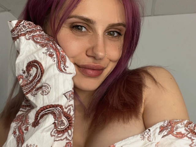 SophieReyX with purple hair, brown eyes, and a nose ring, smiling at the camera, holding a paisley-patterned shirt over her bare boobs. She has a cute, sexy look.