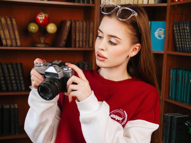 CherylFoxy with long brown hair, wearing a red and white sweater, holding a camera, eyeglasses on her head, looking down, fingers on camera lens, in a library.