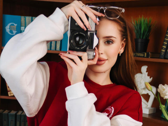 CherylFoxy with red and white sweater, holding vintage camera, eyeglasses on head, smiling, brown hair, subtle makeup, wooden bookshelf, white statue, green plant in background.