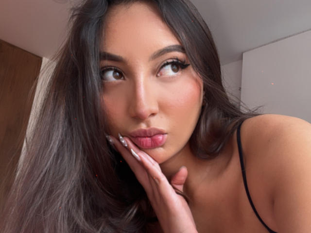 DianaFernandezz with pouty lips, dark brown eyes, long black hair, and a hand touching her cheek, wearing a black spaghetti strap top, looking slightly to the side.