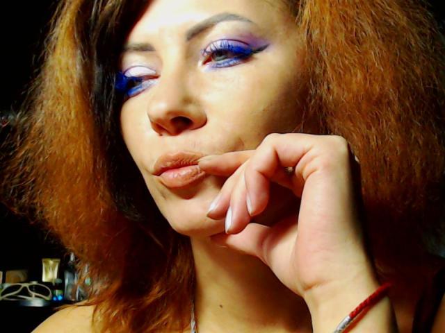 JuliaIce with blue eyeshadow, pouty lips, and wavy brown hair, touching her chin with a finger, wearing a red bracelet, and winking. Her tits are not visible.