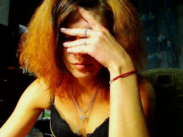 JuliaIce with messy orange-brown hair, black lace top, silver necklaces, and ring, covering face with hand, showing a red bracelet, and a ring on her finger.