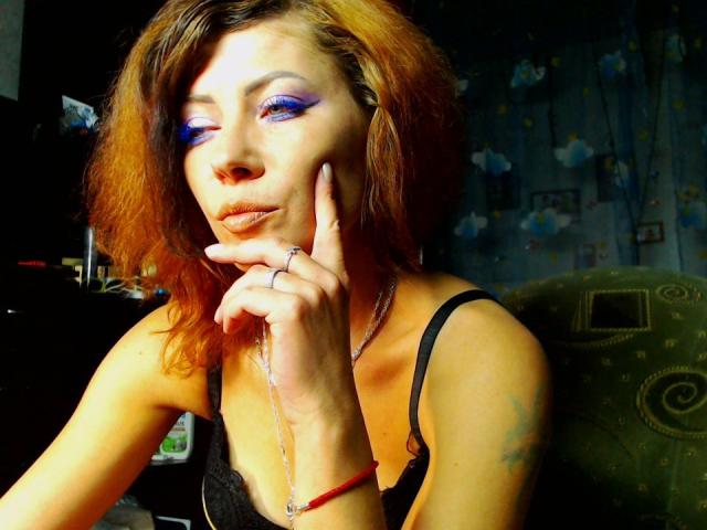 JuliaIce with blue eyeliner, orange hair, black top, finger on chin, ring, necklace, tattoo on arm, green couch, red bracelet, subtle smile, sexy, confident.