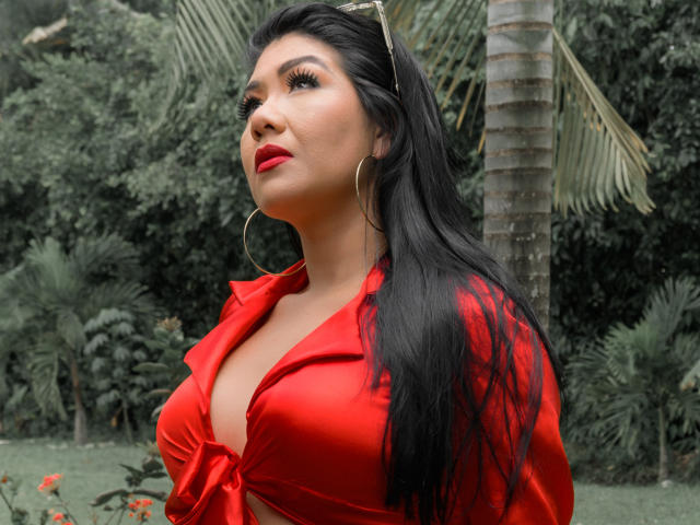 Khloe Lawson, con cabello negro largo, lápiz labial rojo y una camisa roja atada, mostrando sus grandes senos, con aros grandes, de pie frente a una palmera..