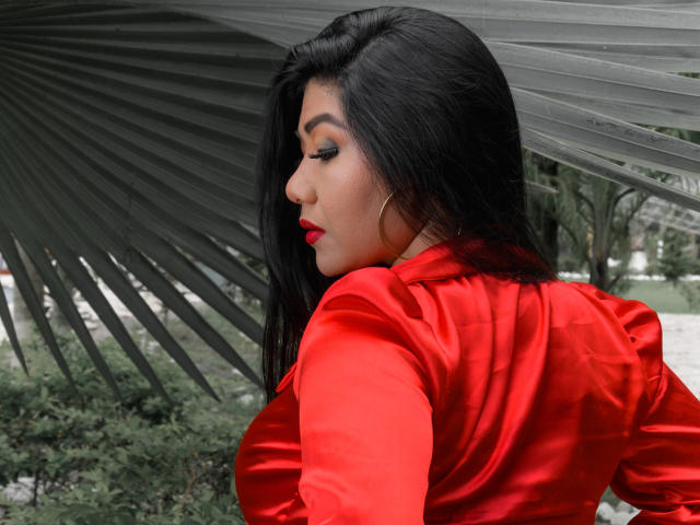 Khloe Lawson, con cabello negro largo, una camisa de satén roja y lápiz labial rojo, mirando hacia un lado, debajo de una gran hoja de palma gris, mostrando su espalda y trasero atractivos..