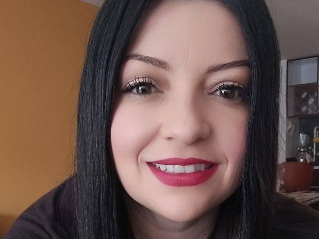 NatalyHami, met recht zwart haar, roze lippenstift en witte tanden, lacht naar de camera, draagt zwarte eyeliner en mascara, en heeft een lichte huidskleur..