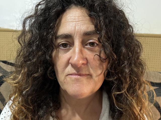 TuFavoritaMilf has curly dark brown hair, a slightly wrinkled face, and a neutral expression. She's wearing a white shirt with small black polka dots.