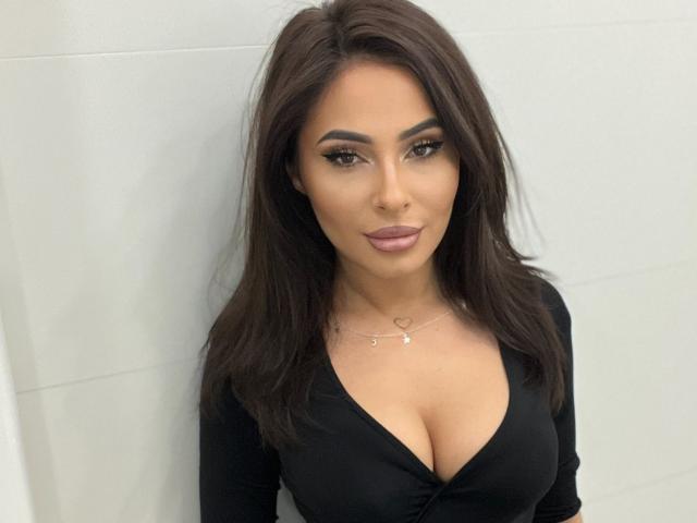 SarahSkye has brown hair, tan skin, and is wearing a black top with a deep cleavage showing her large boobs. She has dark eyeliner and pink lipstick.