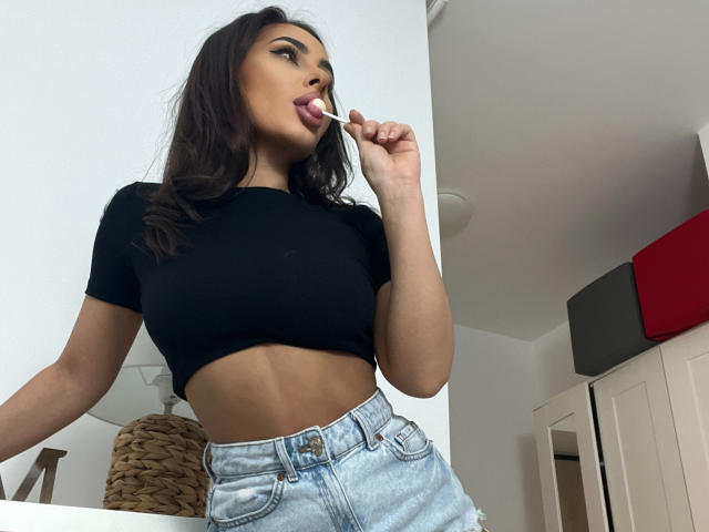 SarahSkye with dark brown hair, black crop top, showing her abs, licking a lollipop, wearing light blue jeans, standing in a kitchen. Her tits are small, her ass is unseen.