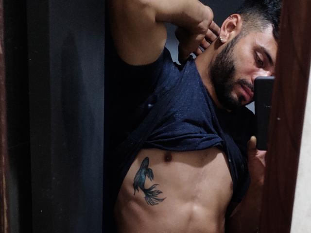JeffSexy lifting his navy shirt, revealing a toned abs with a blue bird tattoo on his right side, short dark hair, and a serious expression in a mirror selfie.