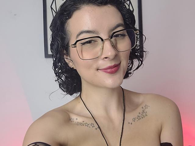 GabrielaMoore with curly black hair, glasses, and red lipstick, smiling with bare shoulders showing star tattoos across chest. Necklace and black off-shoulder top. Sheer, sexy, cute.