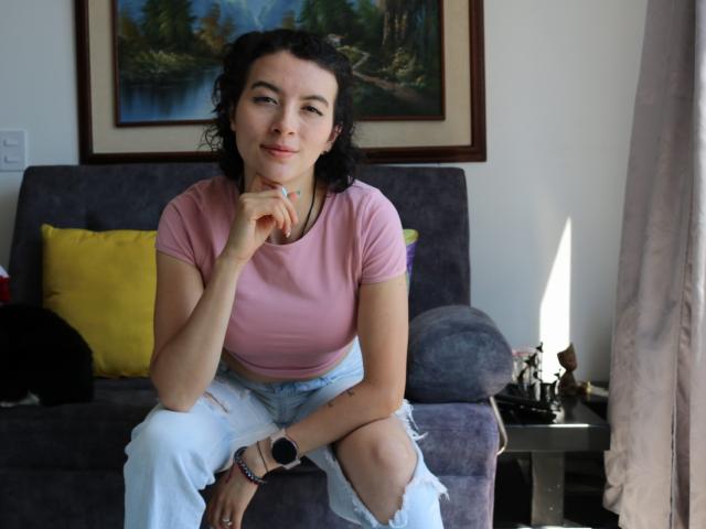 GabrielaMoore with curly black hair, pink tee, and ripped white pants, sitting on gray couch, hand on chin, black watch on wrist, yellow pillow behind, cat on left.